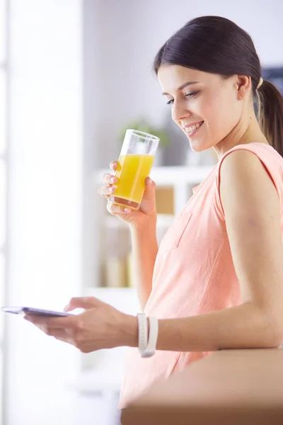 Улыбающаяся красивая женщина смотрит на мобильный телефон и держит стакан апельсинового сока во время завтрака на кухне . — стоковое фото