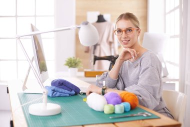 Mutlu moda tasarımcısı gülümsüyor, modern ofis stüdyosunda materyal, kumaş, iplik, tekstil, dizüstü bilgisayarla oturuyor..