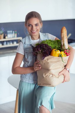 Sebzeli alışveriş poşeti taşıyan genç bir kadın. Mutfakta duruyor.