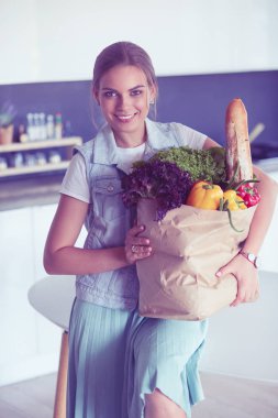 Sebzeli alışveriş poşeti taşıyan genç bir kadın. Mutfakta duruyor.