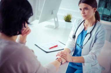 Kadın doktor hastane danışma odasında bir hastayı sakinleştiriyor, elini tutuyor.