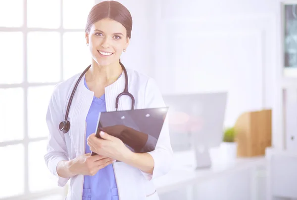 Joven doctora sonriente con estetoscopio sosteniendo una carpeta en una sala de consulta de hospitales — Foto de Stock