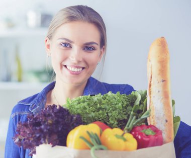 Sebzeli alışveriş poşeti taşıyan genç bir kadın. Mutfakta duruyor.