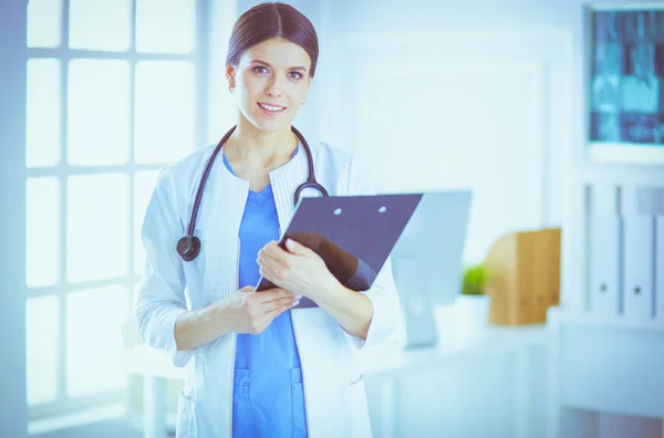 Jonge glimlachende vrouwelijke arts met stethoscoop die een map vasthoudt in een consultatieruimte van een ziekenhuis — Stockfoto