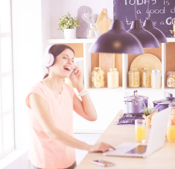 Retrato de una joven alegre escuchando música con auriculares y usando un ordenador portátil mientras está de pie en la cocina — Foto de Stock