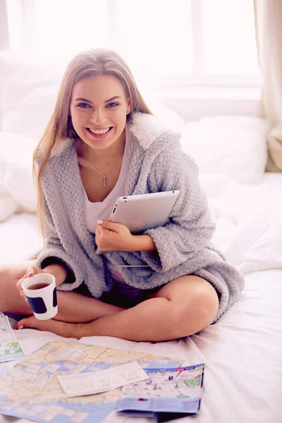 リラックスした若い女性がベッドの上にコーヒーとデジタルタブレットのカップで座っています — ストック写真