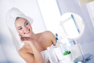 Bornozlu genç kadın banyo aynasına bakıyor.