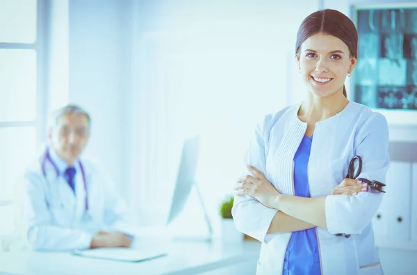 Egy orvos mosolyog a kamerába a férfi kollégájával a kórház rendelőjében. — Stock Fotó