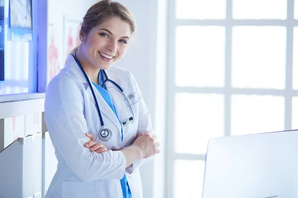 Νεαρή γυναίκα γιατρός με λευκή στολή στέκεται στο γραφείο κλινικές — Φωτογραφία Αρχείου