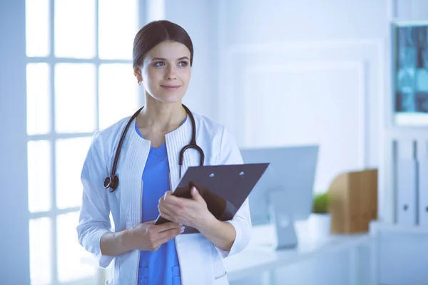 Fiatal mosolygós női orvos sztetoszkóppal kezében egy mappát az orvosok irodájában — Stock Fotó