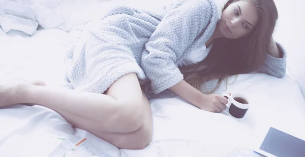Χαλαρός νεαρή γυναίκα κάθεται στο κρεβάτι με ένα φλιτζάνι καφέ και ψηφιακό δισκίο — Φωτογραφία Αρχείου
