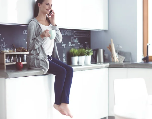 Kobieta przy użyciu telefonu komórkowego siedzi w nowoczesnej kuchni. — Zdjęcie stockowe