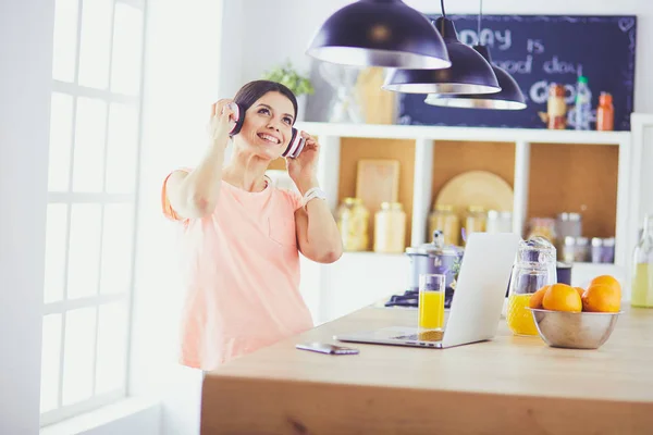 Portrait of a cheerful young woman listening to music with headphones and using laptop computer while standing at the kitchen — Stock Photo, Image