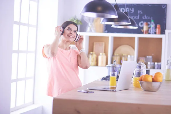 Retrato de una joven alegre escuchando música con auriculares y usando un ordenador portátil mientras está de pie en la cocina — Foto de Stock