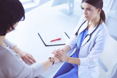 Kadın doktor hastane danışma odasında bir hastayı sakinleştiriyor, elini tutuyor.