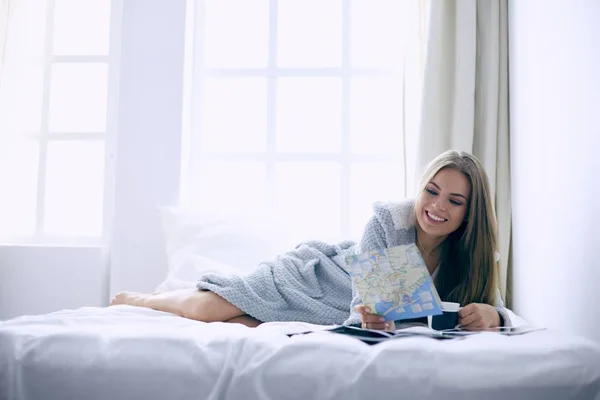 Χαλαρός νεαρή γυναίκα κάθεται στο κρεβάτι με ένα φλιτζάνι καφέ και ψηφιακό δισκίο — Φωτογραφία Αρχείου
