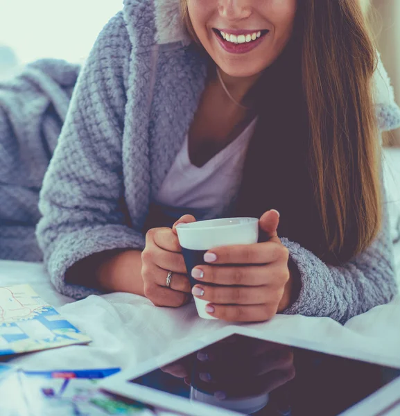 Relaxado jovem mulher sentada na cama com uma xícara de café e tablet digital — Fotografia de Stock