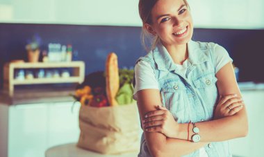 Sebzeli alışveriş poşeti taşıyan genç bir kadın. Mutfakta duruyor.