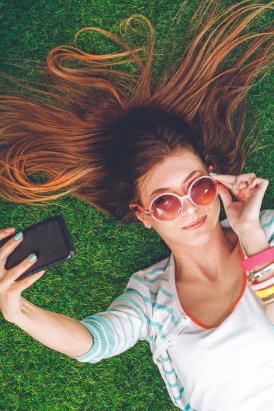 Mooie jonge vrouw die selfie maakt door haar telefoon terwijl ze in groen gras ligt. mooie jonge vrouw maken selfie — Stockfoto