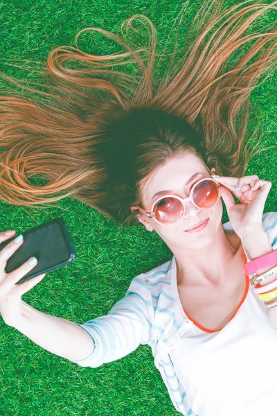 Mooie jonge vrouw die selfie maakt door haar telefoon terwijl ze in groen gras ligt. mooie jonge vrouw maken selfie — Stockfoto