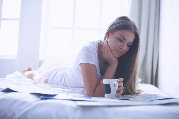 Avslappnad ung kvinna sitter på sängen med en kopp kaffe och digital tablett — Stockfoto