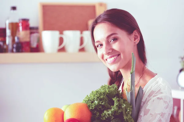 Mujer joven sonriente sosteniendo verduras de pie en la cocina. Jovencita sonriente —  Fotos de Stock