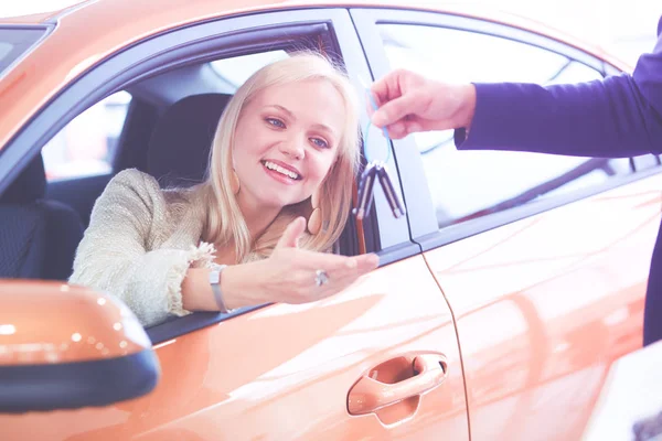 Beautiful young woman is getting key and smiling while sitting in a new car in dealership