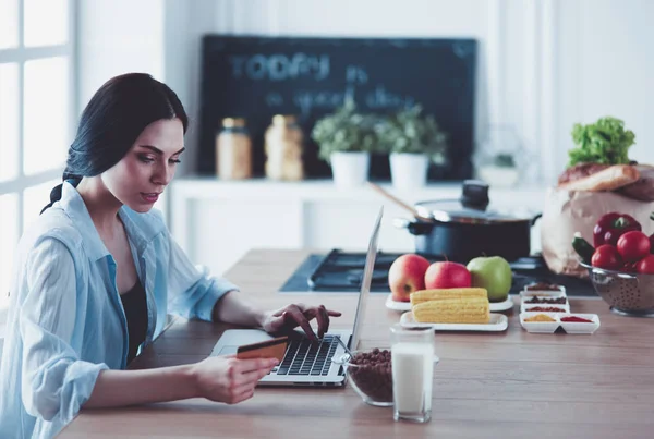 Femme souriante achats en ligne à l'aide d'un ordinateur et carte de crédit dans la cuisine — Photo