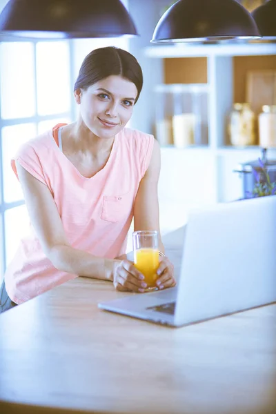Νεαρή γυναίκα στην κουζίνα με φορητό υπολογιστή ψάχνει συνταγές, χαμογελώντας. Έννοια μπλόγκερ τροφίμων — Φωτογραφία Αρχείου