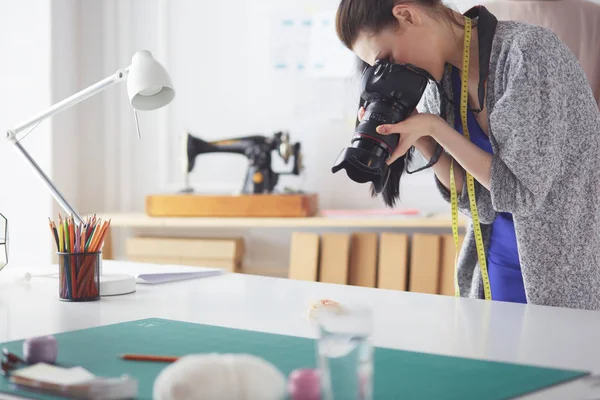 Joven diseñadora tomando fotos con cámara digital mientras está de pie cerca del escritorio en su taller — Foto de Stock