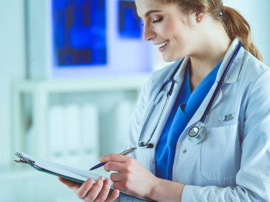 Steteskoplu bir doktor elinde bir defter tutuyor. Hastanede ayakta dikilirken panoda tıbbi form dolduran bir kadın doktorun yakın çekimi.