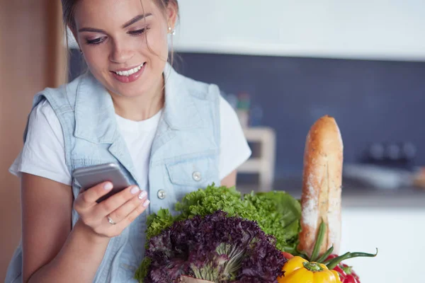 Smiling woman with mobile phone holding shopping bag in kitchen — Stock Photo, Image