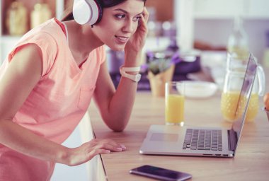 Mutfakta dururken kulaklıkla müzik dinleyen ve dizüstü bilgisayar kullanan neşeli bir genç kadının portresi