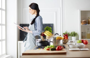Mutfakta yemek yapmak için tablet bilgisayar kullanan genç bir kadın.