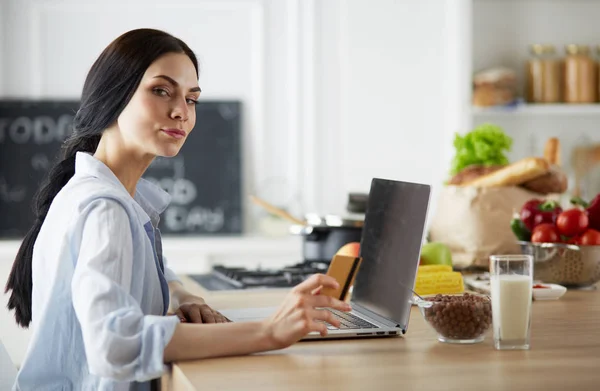 Mujer sonriente compras en línea utilizando la computadora y la tarjeta de crédito en la cocina —  Fotos de Stock