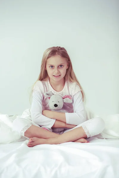 La niña en el dormitorio está sentada en la cama. La niña está usando un pijama y sentada en la cama  . — Foto de Stock