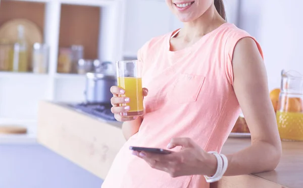 Mujer bonita sonriente mirando el teléfono móvil y sosteniendo un vaso de jugo de naranja mientras desayunaba en una cocina . — Foto de Stock