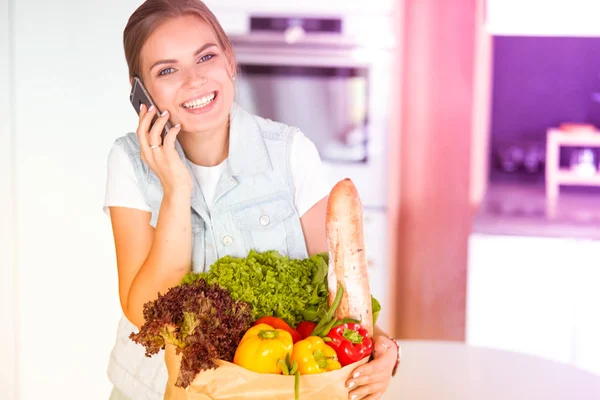 Улыбающаяся женщина с мобильным телефоном держит сумку для покупок на кухне — стоковое фото