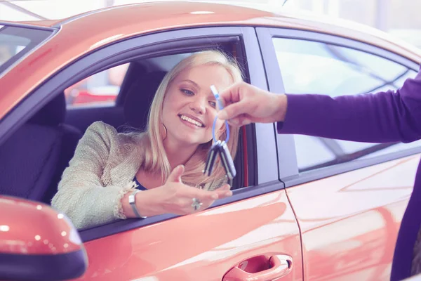 Όμορφη νεαρή γυναίκα παίρνει το κλειδί και χαμογελώντας, ενώ κάθεται σε ένα νέο αυτοκίνητο στην αντιπροσωπεία — Φωτογραφία Αρχείου