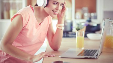 Mutfakta dururken kulaklıkla müzik dinleyen ve dizüstü bilgisayar kullanan neşeli bir genç kadının portresi