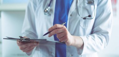 Steteskoplu bir doktor elinde bir defter tutuyor. Hastanede ayakta dikilirken panoda tıbbi form dolduran bir kadın doktorun yakın çekimi.