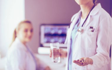 Doktorun arka planda meslektaşıyla klinikte elinde haplar ve bir bardak su tutarken görüntüsü..