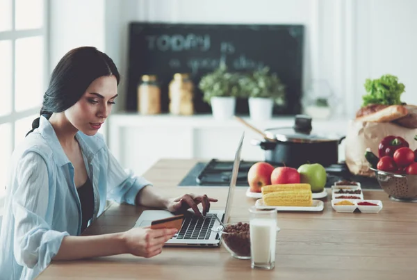 Χαμογελαστή γυναίκα online ψώνια χρησιμοποιώντας υπολογιστή και πιστωτική κάρτα στην κουζίνα — Φωτογραφία Αρχείου