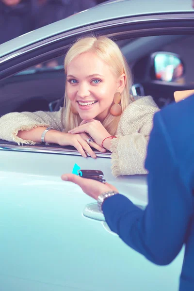 Beautiful young woman is getting key and smiling while sitting in a new car in dealership