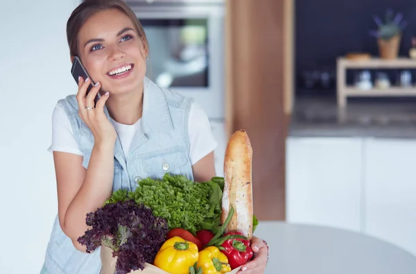 Smiling woman with mobile phone holding shopping bag in kitchen — Stock Photo, Image