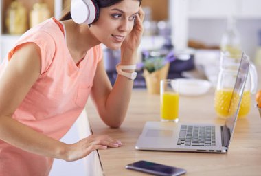 Mutfakta dururken kulaklıkla müzik dinleyen ve dizüstü bilgisayar kullanan neşeli bir genç kadının portresi