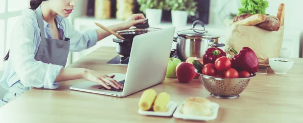 Giovane donna in cucina con computer portatile alla ricerca di ricette, sorridente. Concetto di food blogger — Foto Stock