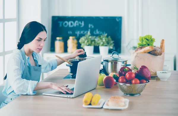 Giovane donna in cucina con computer portatile alla ricerca di ricette, sorridente. Concetto di food blogger — Foto Stock