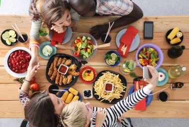Ahşap masada otururken birlikte yemek yiyen bir grup insanın üst düzey görüntüsü. Masada yemek var. İnsanlar fast food yiyor..
