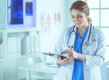 Steteskoplu bir doktor elinde bir defter tutuyor. Hastanede ayakta dikilirken panoda tıbbi form dolduran bir kadın doktorun yakın çekimi.
